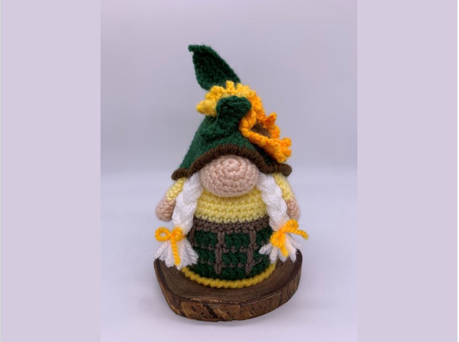 Crocheted amigurumi gnome with dark green pointed hat, sunflower accent, yellow body, green skirt and white braided pigtails on a wood slice.