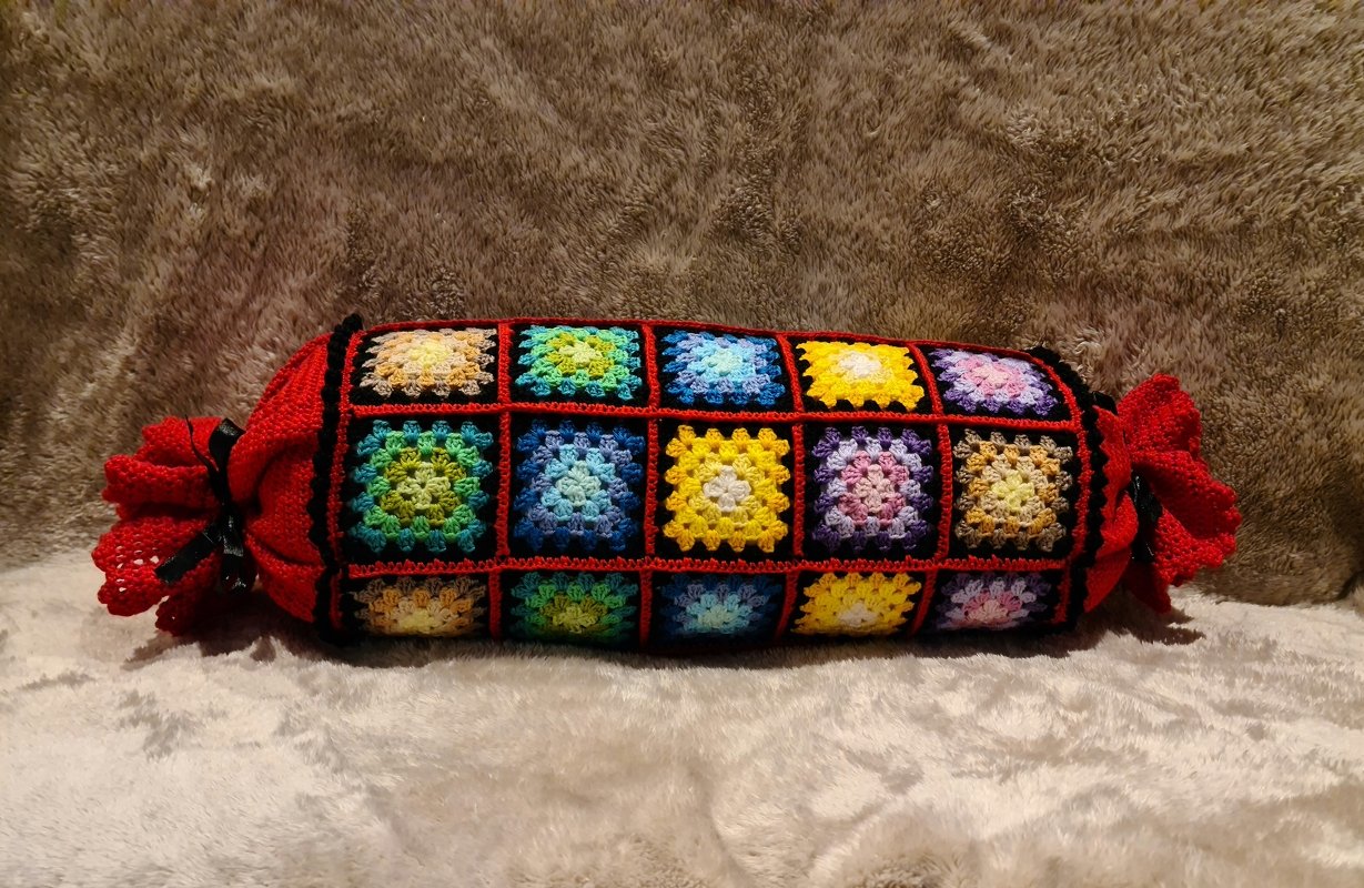 Handgehäkelter Nackenrollenbezug aus bunten Granny-Square-Quadraten mit schwarzer Einfassung und roten gerafften Enden