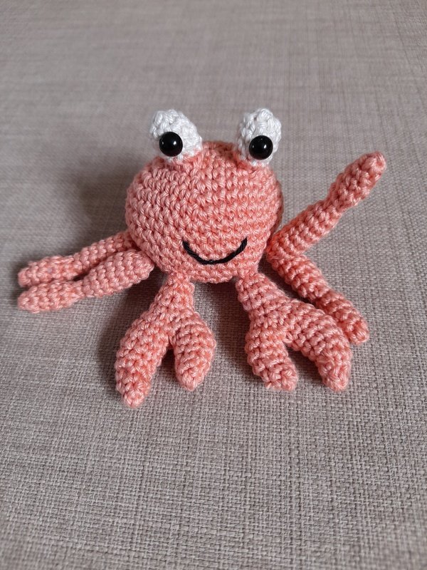 Gehäkelte zartrosa Krabbe mit weißen Augen und gesticktem Lächeln auf hellem Stoff