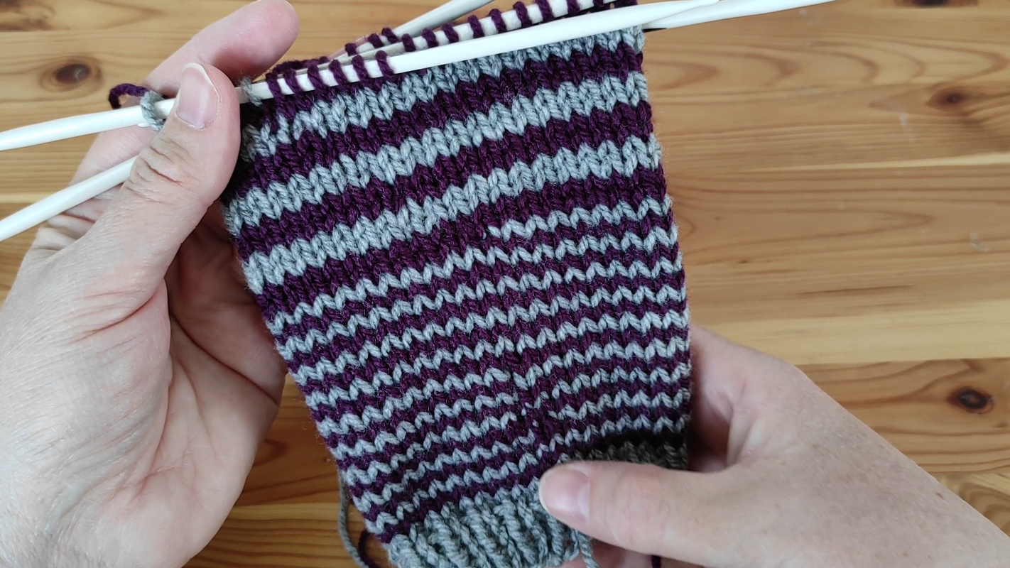 Hände halten ein lila-graues Streifenstrickstück auf weißen Stricknadeln vor einem Holztisch.