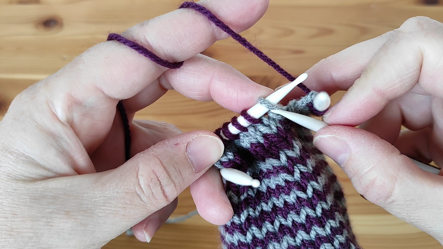 Nahaufnahme von Händen beim Stricken mit Doppelspitzen, mit violetter und grauer Wolle