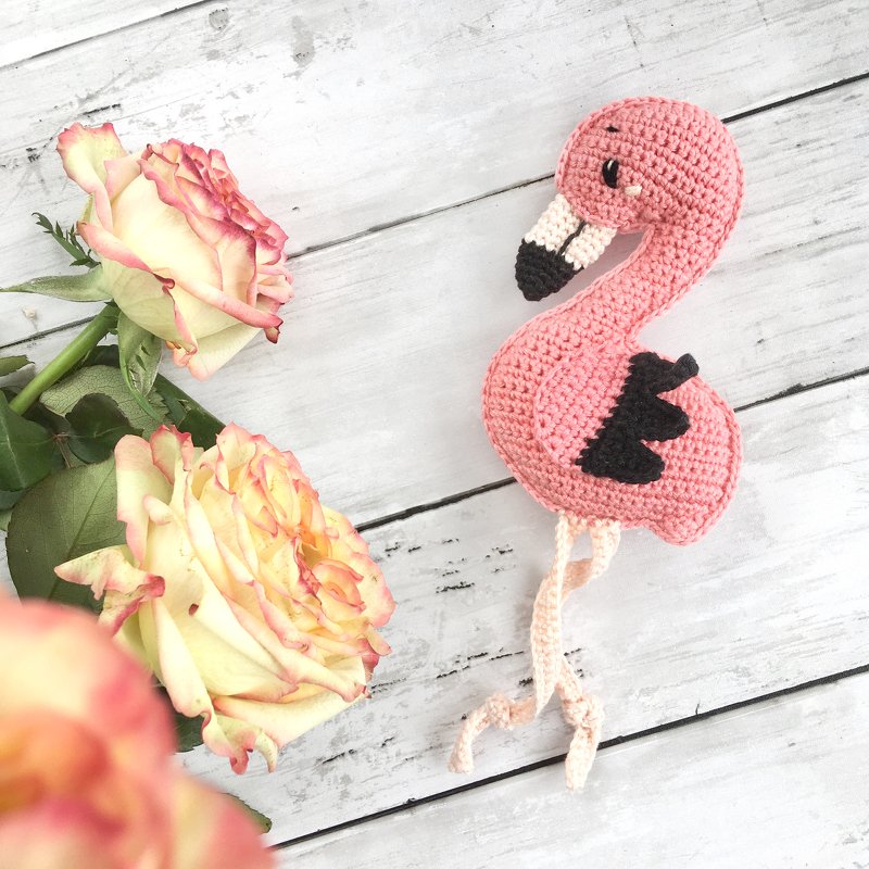 Nahaufnahme eines gehäkelten rosa Flamingos neben zwei rosafarbenen Rosen auf hellem Holz.