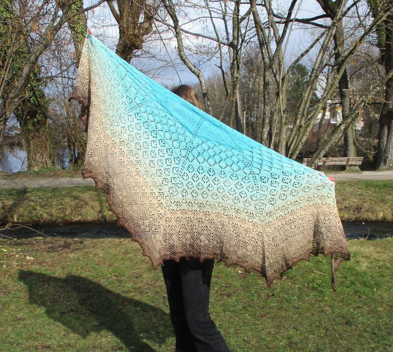 Person hält dreieckiges Lacetuch mit türkis‑beige‑braunem Ombré und Lochmuster im Freien hoch