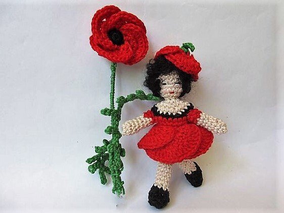 Gehäkelte Amigurumi-Puppe mit schwarzem Haar im roten Kleid und rotem Hütchen, daneben eine große rote Mohnblume am grünen Stiel.