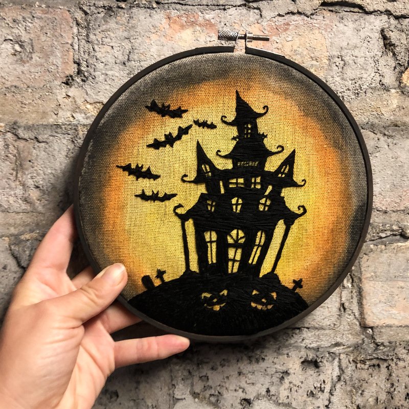 Hand hält runden Stickrahmen mit Halloween-Stickbild: schwarze Spukhaus-Silhouette, Fledermäuse und Kürbisse vor orange-gelbem Hintergrund.