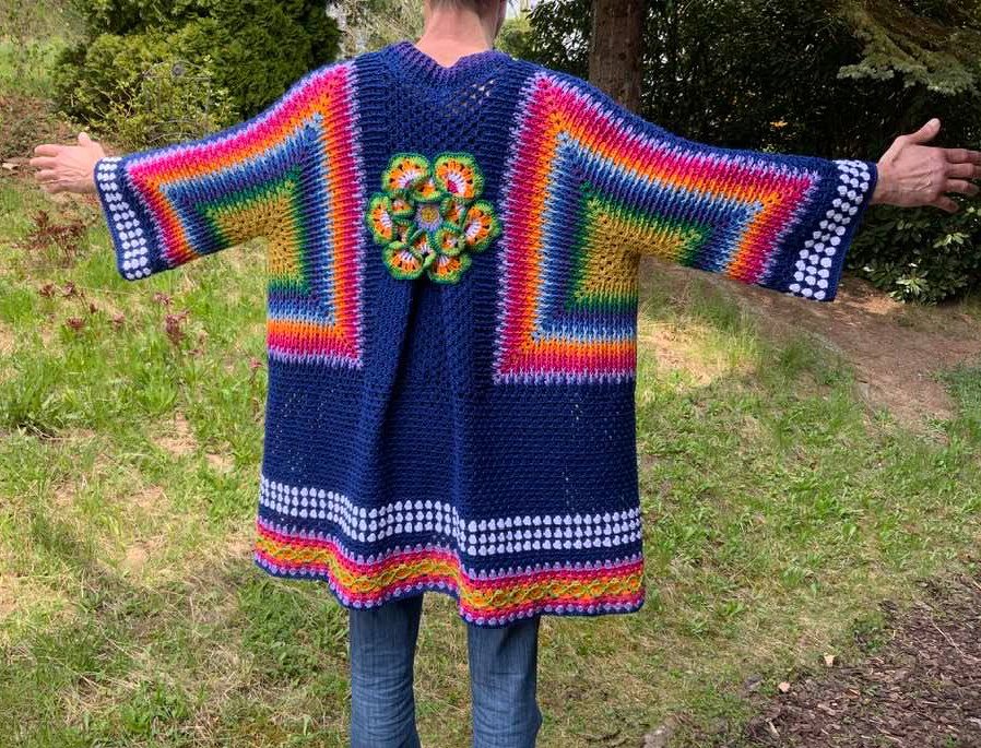 Rückenansicht einer Person in dunkelblauer Häkeljacke mit Regenbogenstreifen an Ärmeln und Saum und einer grünen-orangen Blumenapplikation auf dem Rücken.