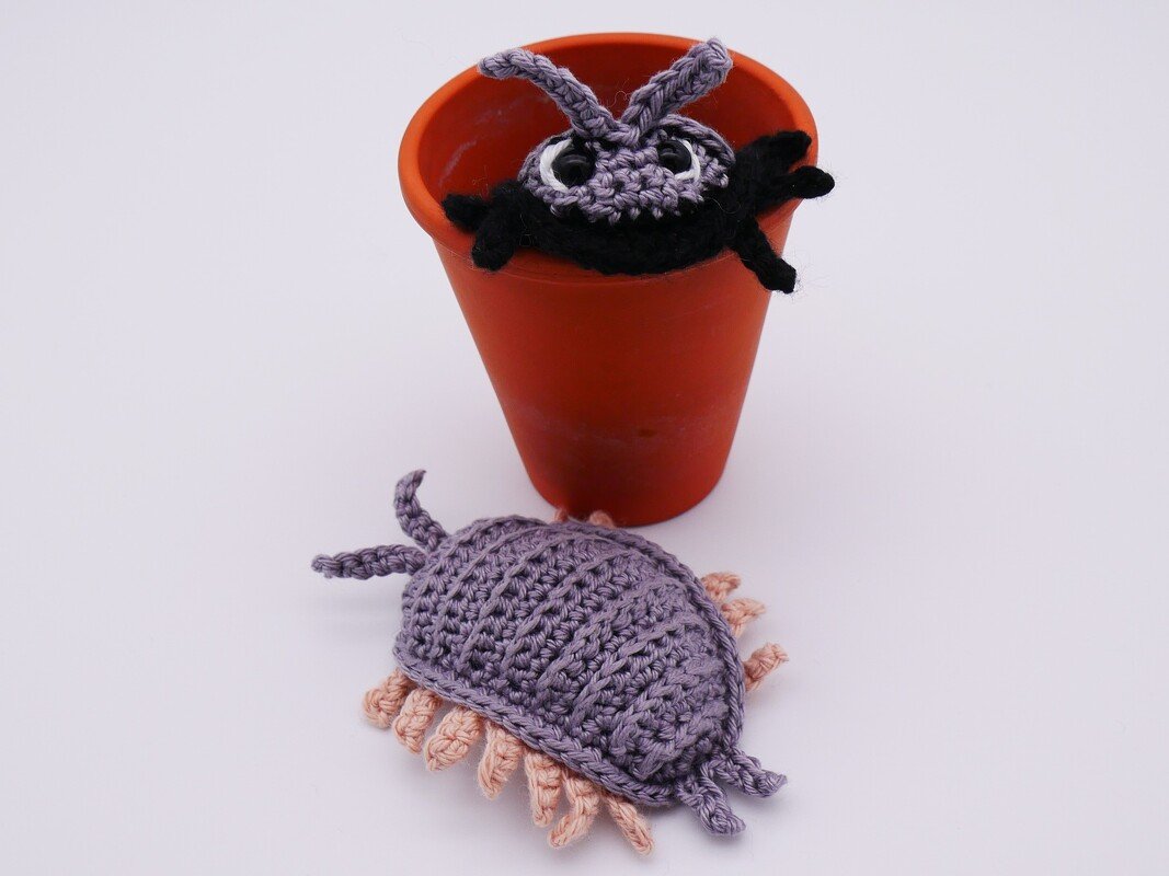 Zwei gehäkelte Mini-Amigurumi: flieder mit rosa Fransen und schwarz-lila in orangefarbenem Topf