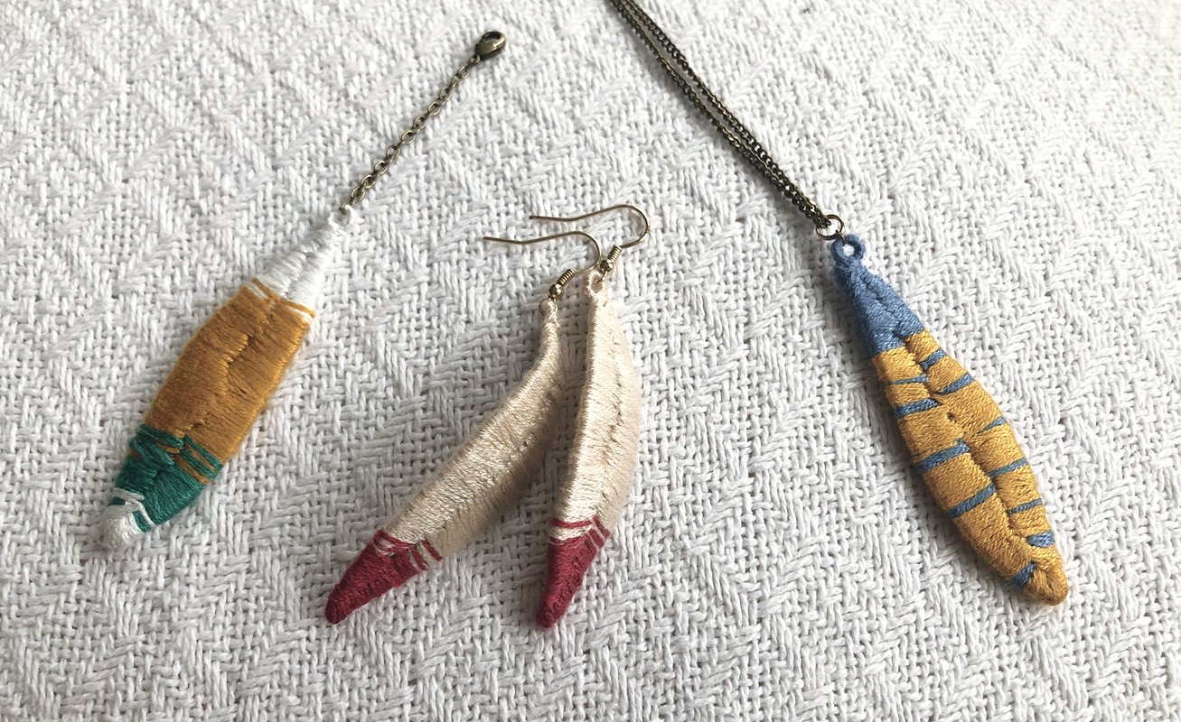 Zwei bestickte Anhänger und ein Paar bestickte Hängeohrringe auf weißem, strukturiertem Stoff; Farben: Senfgelb, Blau, Grün, Beige, Rot.