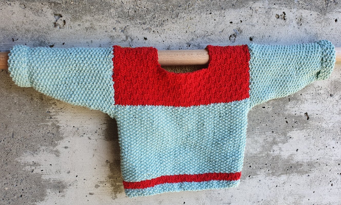 Baby-Pullover in Hellblau mit rotem Einsatz und Saumstreifen, gestrickt im Krausmuster, an einer Holzstange präsentiert.