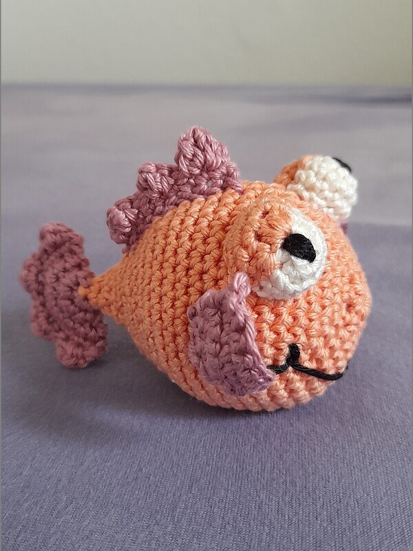 Gehäkelter Amigurumi-Fisch orange mit lila Flossen auf grauem Hintergrund
