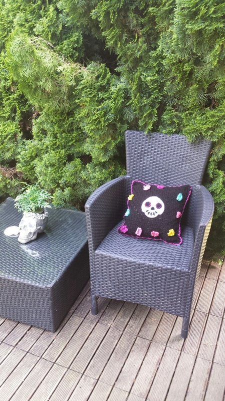 Schwarzes gehäkeltes Kissen mit weißem Totenkopf und bunten Applikationen auf Rattanstuhl