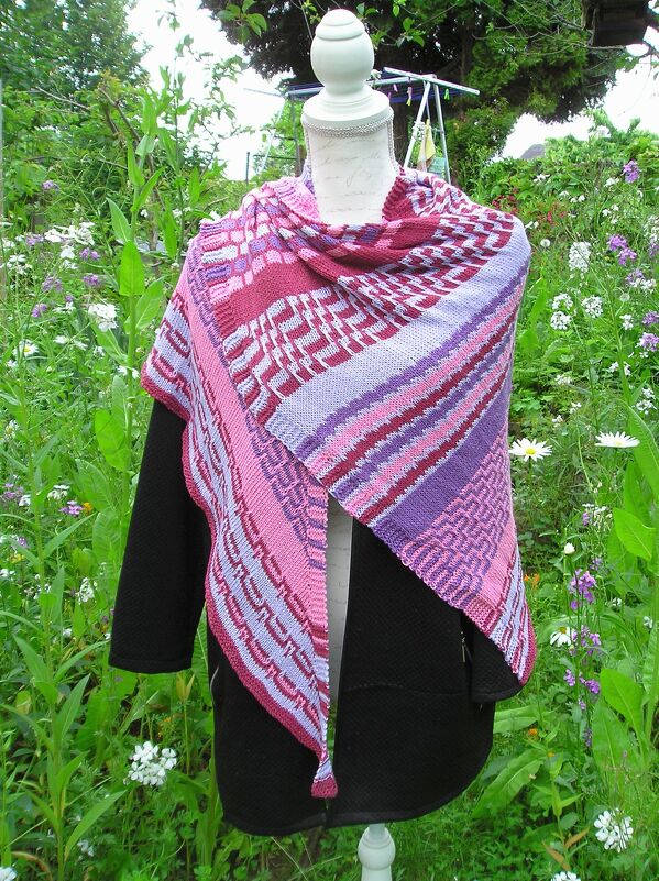 Gestricktes Dreieckstuch in Pink, Lila und Burgunder mit Streifen- und geometrischen Mustern auf einer Schaufensterpuppe vor Gartenpflanzen.