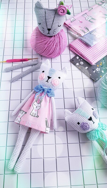Assortment of amigurumi cat parts: crocheted cat heads and legs, balls of pink and mint yarn, a crochet hook and folded fabric panels with bunny prints on a gridded surface.