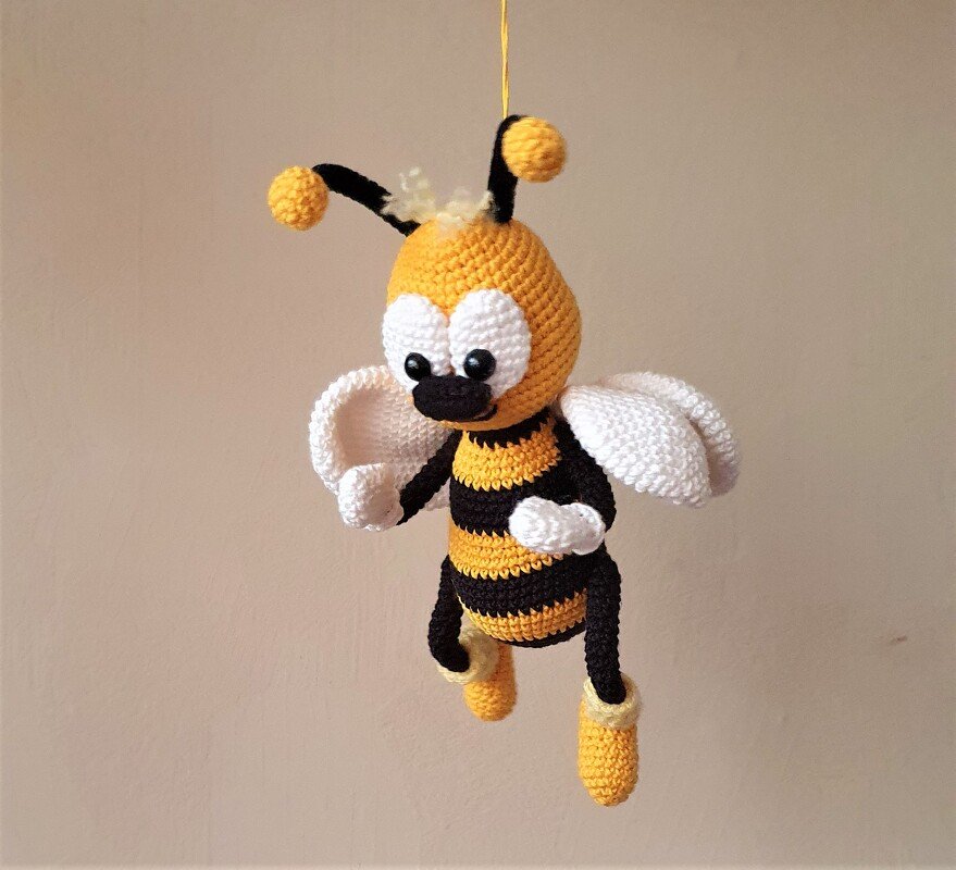 Gehäkelte Amigurumi-Biene, gelb-schwarz gestreift, mit großen weißen Augen und Flügeln an einem gelben Faden hängend.