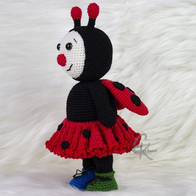 Gehäkeltes Marienkäfer-Amigurumi mit rotem Rock, schwarzen Punkten, Flügeln und bunten Schuhen auf weißem Hintergrund