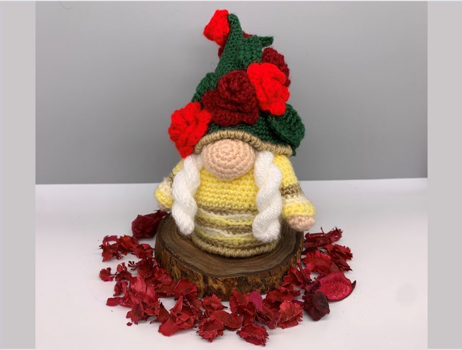 Gehäkelter Amigurumi‑Wichtel mit grünem Blumenhut, gelb gestreiftem Körper und weißen Zöpfen auf einer Baumscheibe, umgeben von roten Blüten.