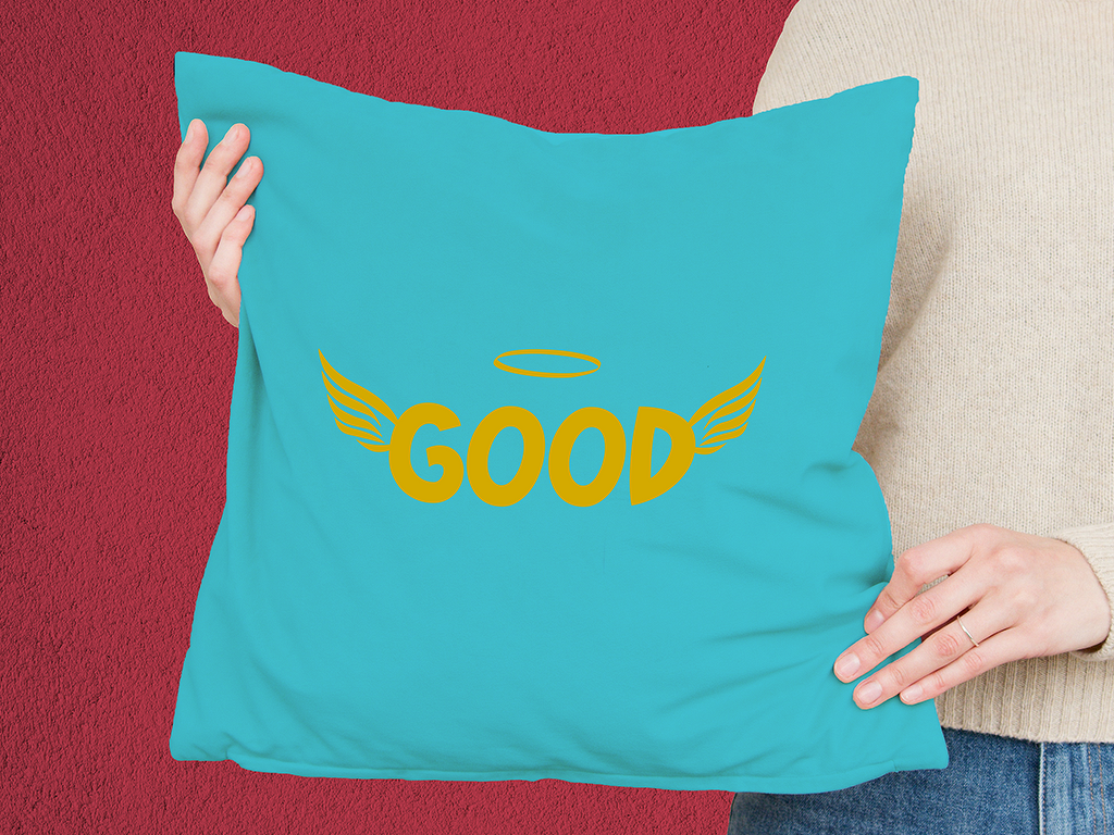 Türkisfarbenes Kissen mit gelbem Schriftzug „GOOD“, stilisierte Flügel und Heiligenschein, gehalten von Händen