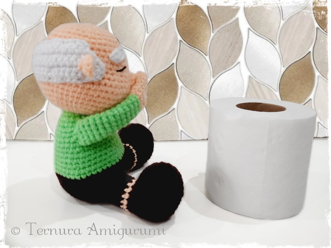 Gehäkelte sitzende Amigurumi-Puppe mit grünem Pullover neben einer Toilettenpapierrolle