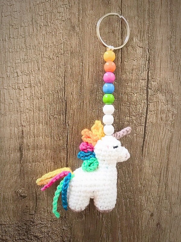 Kleiner gehäkelter Einhorn-Schlüsselanhänger mit regenbogenfarbener Mähne und bunten Perlen am Schlüsselring vor Holz