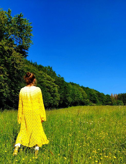 Rückansicht einer Person in langer gehäkelter gelber Jacke auf grüner Wiese vor blauem Himmel