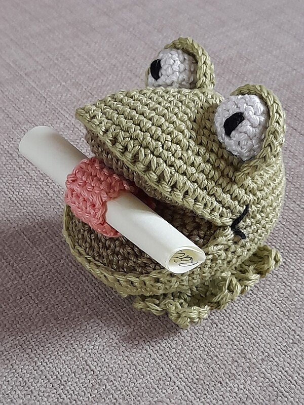 Hellgrüner gehäkelter Frosch-Amigurumi mit rosa Zunge, hält gerollte Notiz
