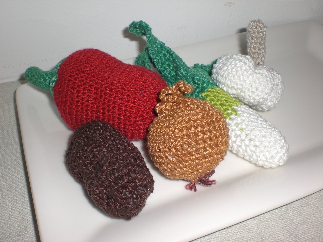 Fünf gehäkelte Gemüsefiguren: Paprika, Zwiebel, Lauch, Kartoffel und Pilz auf weißer Platte.