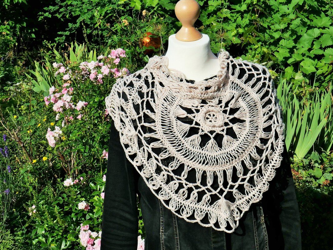 Hellbeiges gehäkeltes Dreieckstuch auf Büste vor grünen Pflanzen und rosa Blumen