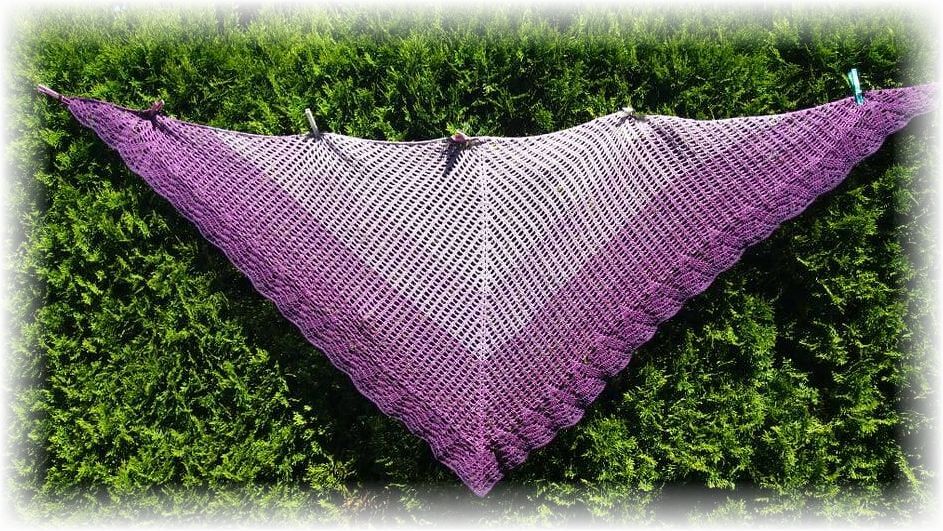 Gehäkeltes lila Dreieckstuch mit Muschelrand vor grüner Hecke aufgehängt