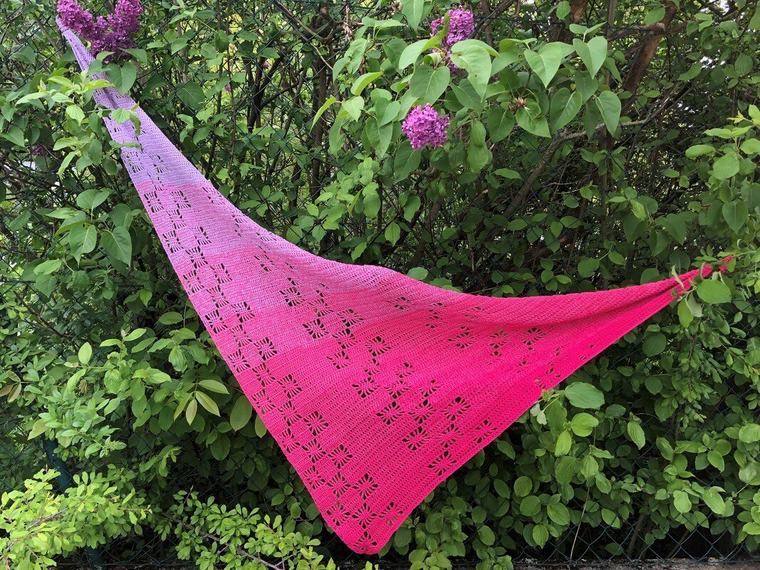 Gehäkeltes Dreieckstuch mit lila-rosa Farbverlauf und quadratischem Lochmuster, aufgehängt an einem Strauch