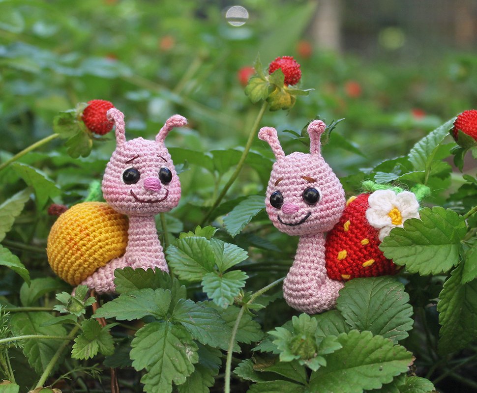 Zwei rosa gehäkelte Amigurumi-Schnecken mit gelbem und Erdbeerhaus zwischen Erdbeerpflanzen