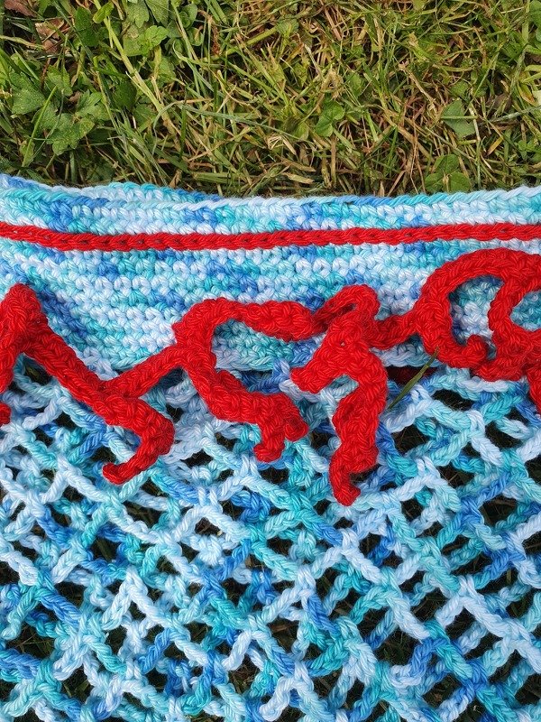 Gehäkelte Netztasche mit blau-türkisfarbenem Netzmuster und roter Zierkordel auf Gras