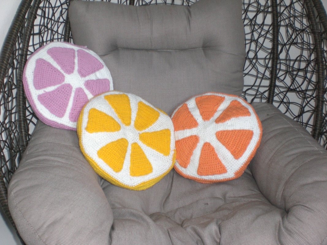 Drei gehäkelte, runde Kissen im Fruchtscheiben-Design (rosa, gelb, orange) auf grauem Hängesessel