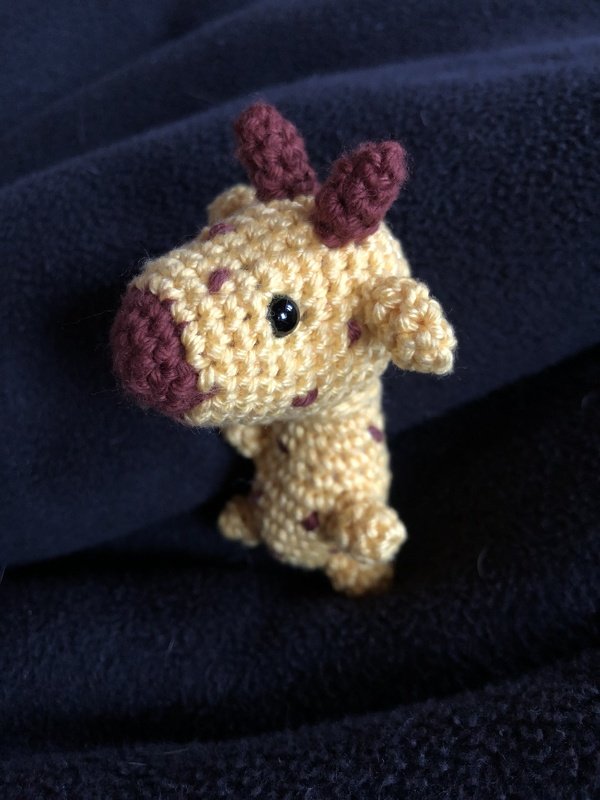 Nahaufnahme einer kleinen gelben Amigurumi‑Giraffe mit braunen Punkten auf dunklem Stoff