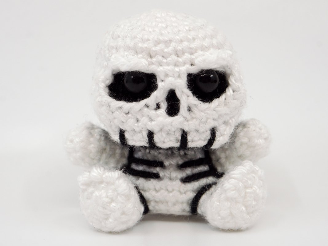 Small white amigurumi crochet skeleton toy with black eyes and stitched ribs on a plain white background.