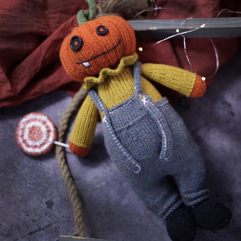 Knitted pumpkin amigurumi doll with grey overalls holding a striped lollipop