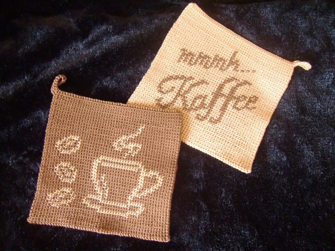 Zwei gehäkelte Topflappen auf dunklem Stoff: hellbeiger mit dem Schriftzug „mmmm... Kaffee“, brauner mit dampfender Kaffeetasse und Kaffeebohnen.