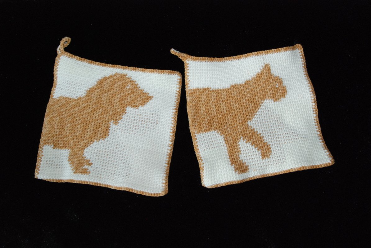 Pair of crocheted square potholders with tan dog and cat silhouettes on white, placed on a black surface.
