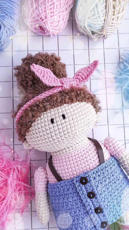 Crocheted amigurumi doll with curly brown hair, pink bunny ear headband and blue overalls on a white grid with yarn skeins.