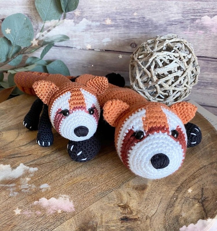 Zwei gehäkelte rote Panda-Schmusetücher mit weißen Gesichtern und schwarzen Pfoten auf Holztisch