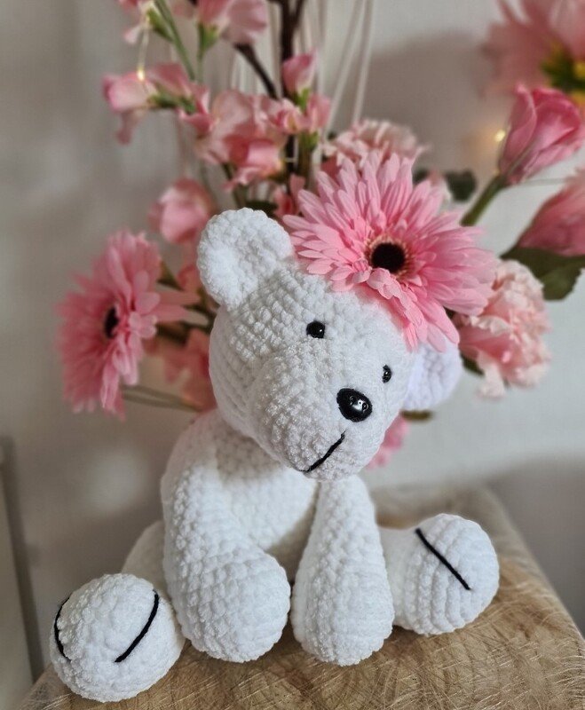 Weißer gehäkelter Amigurumi-Bär mit einer rosa Blüte auf dem Kopf vor rosa Blumen