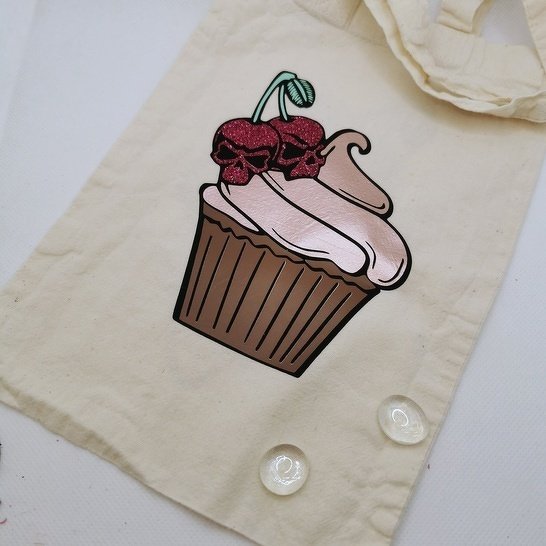 Cupcake-Plottdesign mit rosa Glasur und zwei glitzernden, dunkelroten Totenkopf-Kirschen auf hellem Stoff.