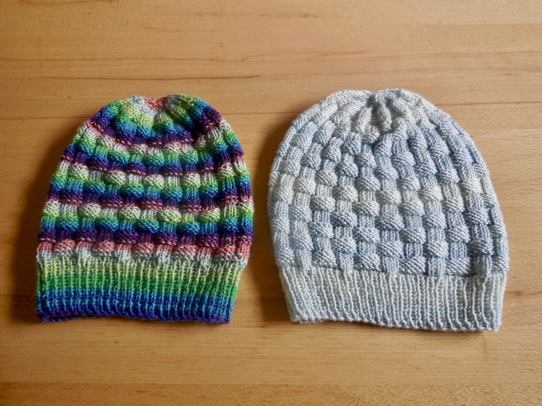 Zwei gestrickte Mützen: eine bunte mit Farbverlauf, eine hellblau-weiße, beide mit texturiertem Muster auf Holzhintergrund.