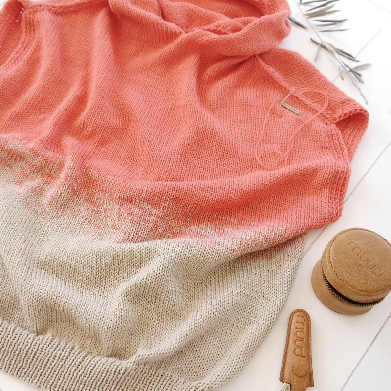 Coral-to-beige ombre knit garment with knitting needle and leather accessories on a white background.