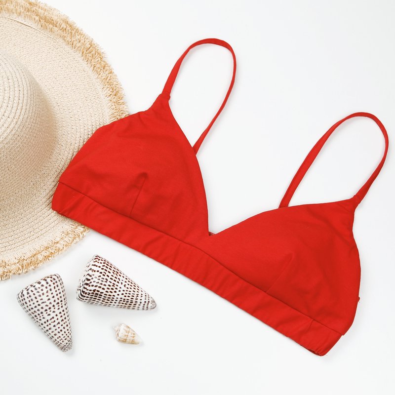 Flach liegendes rotes Bralette-Bikinioberteil mit sichtbarem Unterbrustband, daneben Strohhut und Muscheln