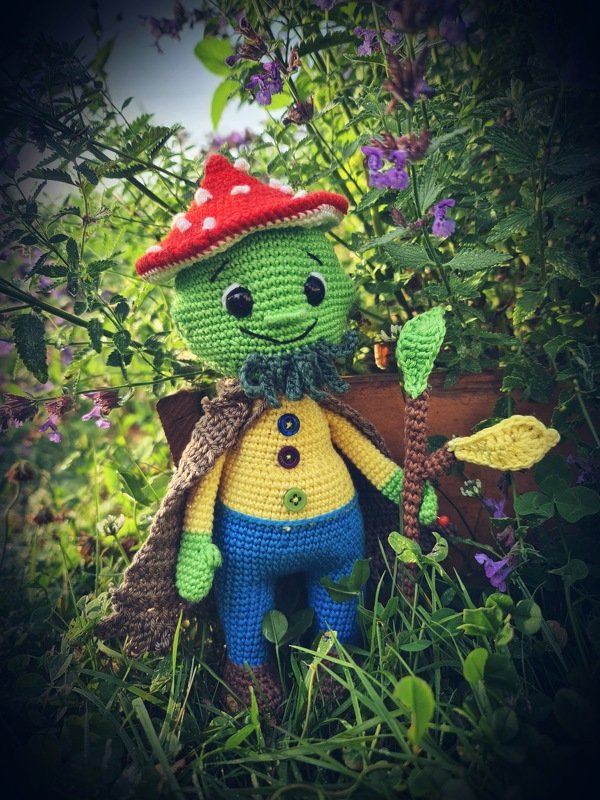 Gehäkelte Amigurumi-Puppe mit rotem Pilzhut, Umhang und Stab in Wiese
