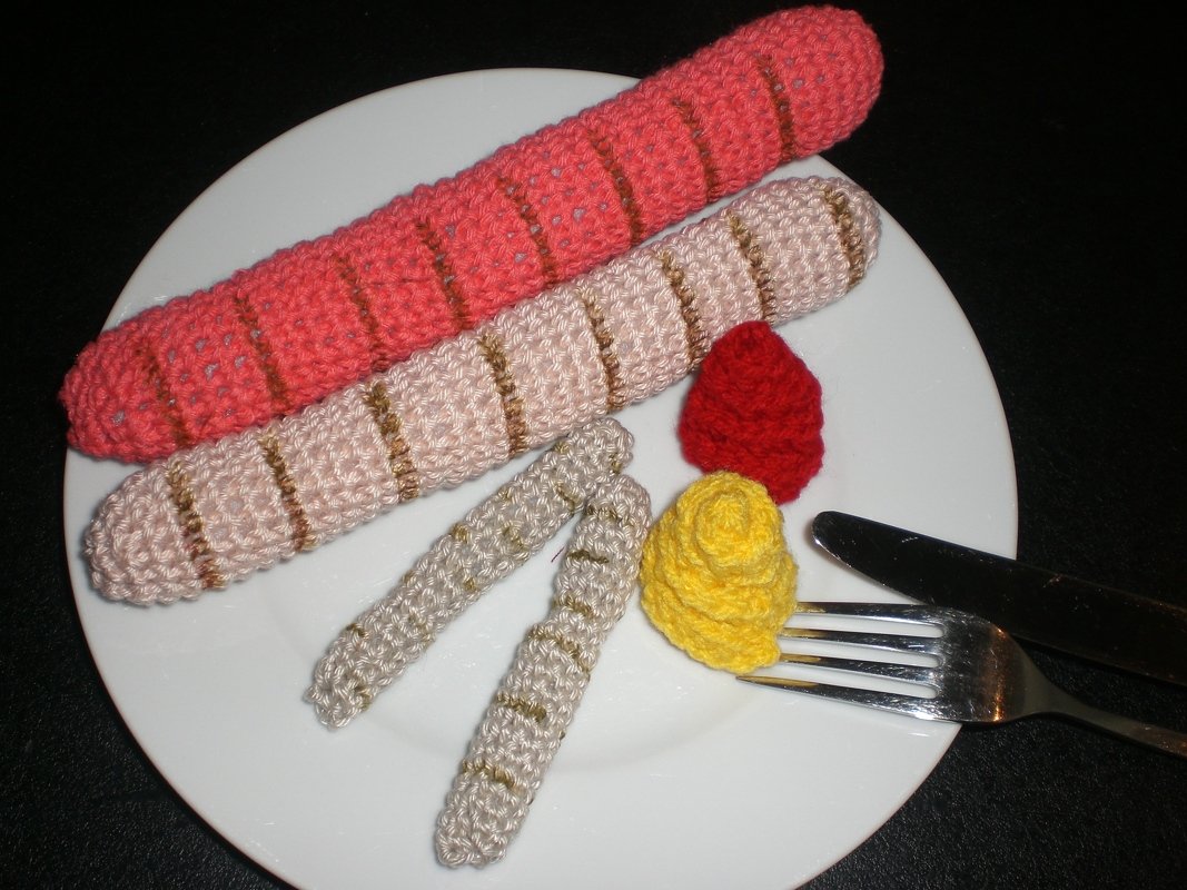 Weißer Teller mit gehäkelten Würstchen, Pommes und roten sowie gelben Soßenklecksen; Besteck auf schwarzem Hintergrund.
