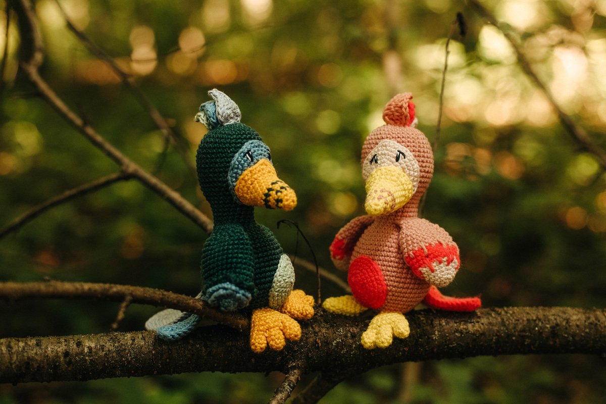 Zwei gehäkelte Amigurumi-Vögel in Grün und Rosa auf einem Ast vor unscharfem, grünem Hintergrund.