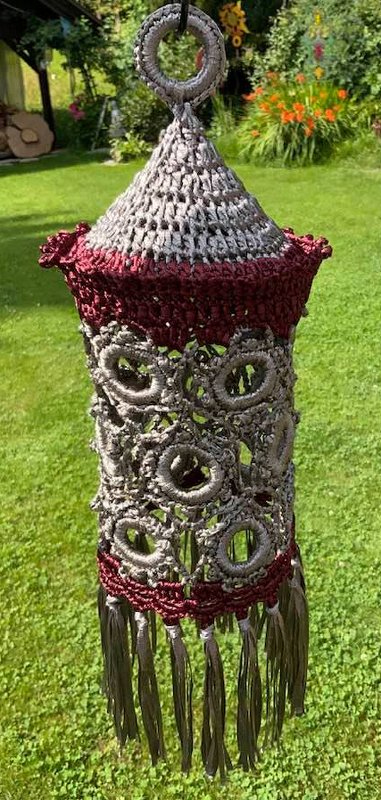 Gehäkelter Lampion in Grau und Dunkelrot mit Ringmustern und Quasten, aufgehängt im Garten.