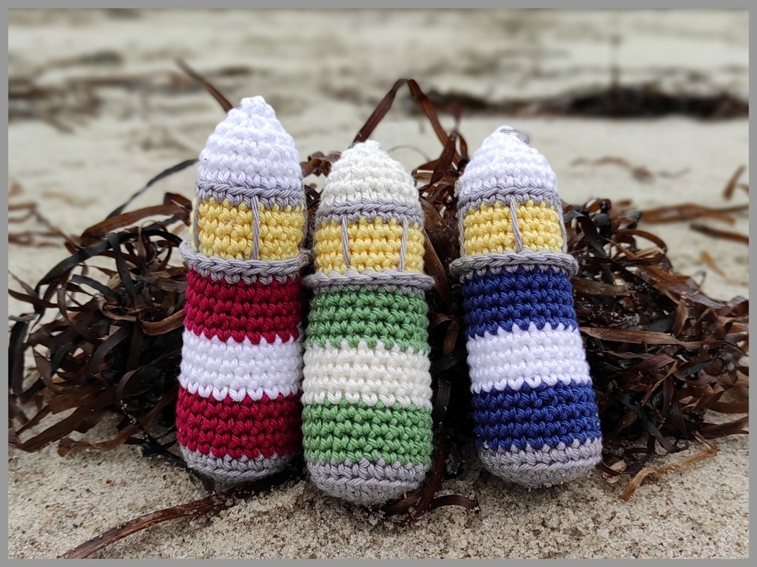 Close-up of three striped crocheted lighthouse toys on sand with seaweed in red, green, and blue.