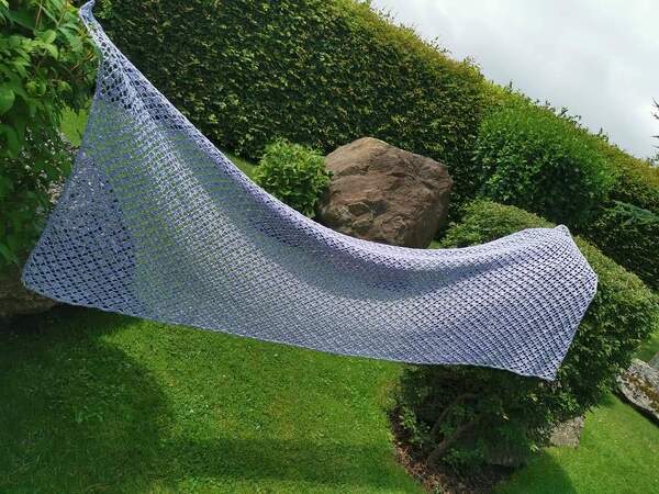 Hellblauer, dreieckiger Häkelschal mit offenem Spitzenmuster, aufgehängt vor grünen Büschen.